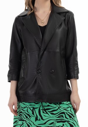 Femme portant un blazer en cuir noir avec double boutons et une jupe verte à rayures zèbre noires, plus des colliers en or superposés.