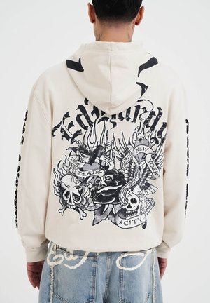 Sudadera color beige con un diseño gráfico que presenta calaveras, rosas y banderas. Impresiones en tinta negra en la parte posterior y las mangas, mostrando una estética inspirada en los tatuajes.