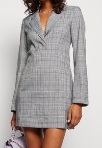 Robe à carreaux gris avec une coupe ajustée, un col en V profond et des manches longues. Le tissu présente des motifs à carreaux avec une texture lisse.