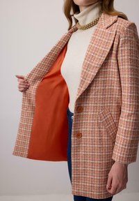 Blazer s oranžovými a smotanovými štvorcami s výstrihom do "V", dvoma hnedými gombíkmi a oranžovou podšívkou; nosený cez krémový rolák.