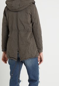 Cazadora parka en verde olivo con capucha, cintura con cordón ajustable y un dobladillo trasero ligeramente más largo. La tela parecetexturizada y resistente.