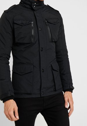 Mann trägt schwarze Jacke mit mehreren Klappen- und Reißverschlusstaschen vorne, schwarzen Knöpfen und hohem Kragen, kombiniert mit schwarzer Hose.