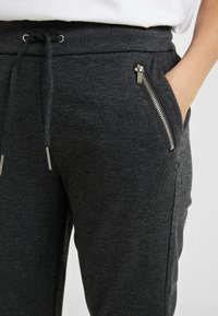 Mörkgrå mjukisbyxor i mjukt tyg, med dragsko i midjan, fickor med dragkedja på sidorna och en slät yta.