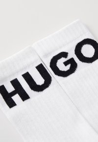 Chaussettes en coton blanches avec des lettres tricotées épaisses et noires "HUGO" sur les bords. La texture est côtelée, avec une finition lisse.