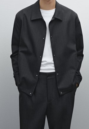 Homme portant une veste boutonnée gris foncé par-dessus un t-shirt blanc et un pantalon gris foncé assorti, debout les mains dans les poches.