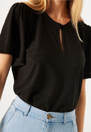Femme portant un chemisier noir à manches courtes en broderie anglaise avec une petite encolure goutte d'eau, rentré dans un jean bleu clair taille haute.
