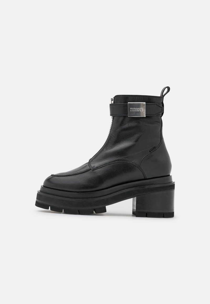 Bottine en cuir noir avec semelle épaisse, talon bloc, fermeture éclair avant et sangle latérale avec boucle en métal marquée.