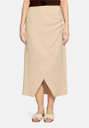 Jupe midi beige portefeuille avec fronces latérales portée avec des sandales slides beige, montrée de la taille aux pieds sur fond uni.