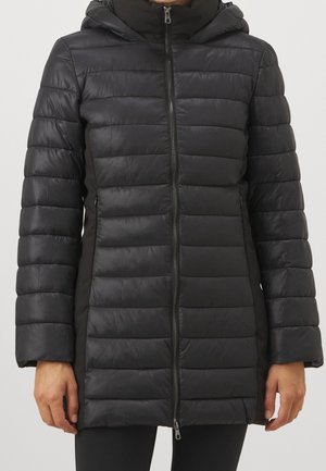 Veste d’hiver noire matelassée avec capuche et fermeture éclair portée par une personne debout, montrée du cou aux hanches avec les mains sur les côtés.