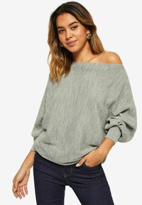 Maglione grigio a coste con design off-shoulder, vestibilità ampia e texture sottile. Abbinato a jeans scuri, che mostra uno stile rilassato.