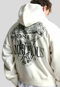 Krém színű kapucnis pulóver, amelynek hátulján nagy fekete grafikai nyomtatás található újságcímekkel és virágos elemekkel. Puha anyagból készült, lazán illeszkedő fazonnal.