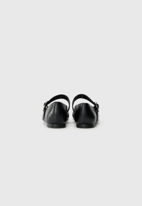 Ballerines en cuir noir avec un bout arrondi, une sangle ajustable et un accent en boucle argentée. Texture lisse et semelle plate. Vue arrière montrée.