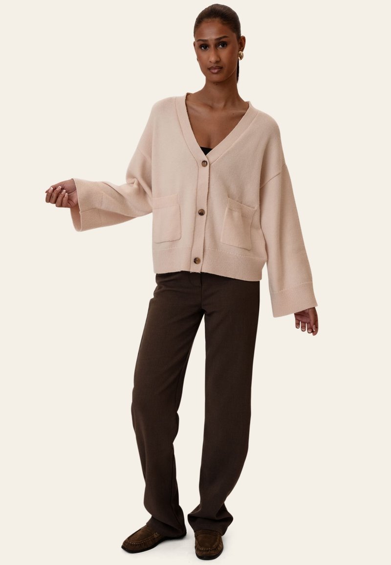 Kvinde iført en beige oversized cardigan med knapper, mørkebrune bukser og brune mokkasinstil sko, stående med armene let udstrakt.