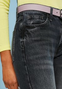 Personne portant un jean noir délavé avec poches avant et poche monnaie, une ceinture rose clair et un haut jaune à manches longues sur fond bleu.