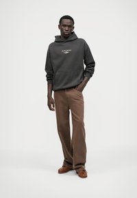 Hallide grei kapuutsiga, millel on eesmine tasku ja logo, koos pruuni velskakanga pükste ja pruuni mokassiiniga. Neutraalne taust rõhutab riietust.