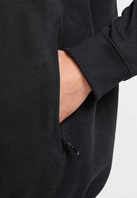 Black fleece jacket with a zippered pocket and a small drawcord. Features a soft texture and ribbed cuffs, designed for comfort and functionality.