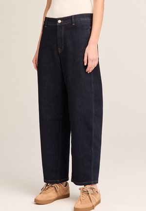 Person wearing dark blue wide-leg jeans with visible orange stitching, beige sneakers, and a white top against a plain beige background.