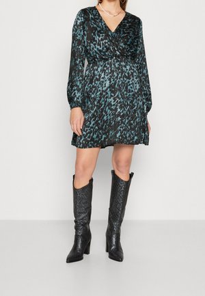 Femme portant une robe à manches longues bleue et noire à motifs avec des bottes noires texturées montantes à talons, sur un fond uni.