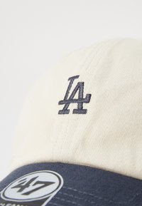Baseballpet met een beige katoenen kroon en een marineblauwe klep, versierd met een geborduurd "LA"-logo in marineblauw en een ronde logotag.