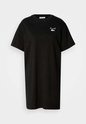 Crna pamučna majica s opuštenim krojem, kratkim rukavima, okruglim izrezom i bijelim Lacoste logotipom na lijevoj strani prsa. Minimalistički dizajn.