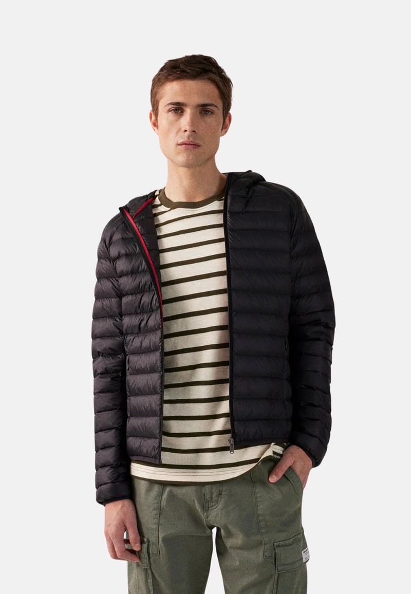 Giovane uomo che indossa una giacca a vento nera sopra una maglietta a righe bianche e verdi e pantaloni cargo verdi, in piedi con una mano in tasca.