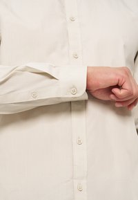 Personne ajustant le poignet d'une chemise beige clair manches longues avec quatre boutons visibles sur le devant et la manche.