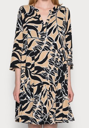 Kvinde iført en knælængde beige kjole med sort og hvidt abstrakt bladmotiv, 3/4 ærmer og en V-udskæring.