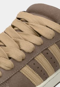 Καφέ πάνινο παπούτσι από σουέτ με μπεζ κορδόνια, που διαθέτει τρεις μπεζ ρίγες, οπές και λευκή λαστιχένια σόλα για αντίθεση.