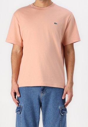 Mężczyzna noszący jednokolorowy jasno różowy t-shirt z krótkim rękawem z małym zielonym logo krokodyla na piersi oraz niebieskie jeansy typu cargo.