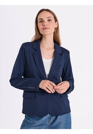 Jonge vrouw met recht lichtbruin haar draagt een marineblauw blazer over een wit topje en blauwe jeans, staand tegen een effen witte achtergrond.