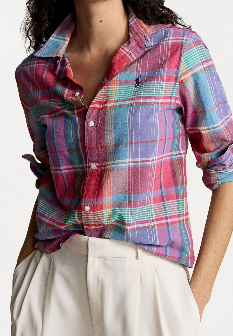 Femme portant une chemise à manches longues à carreaux colorés retroussée, rentrée dans un pantalon crème taille haute, les mains dans les poches.