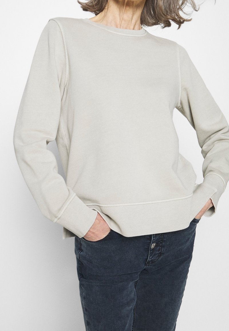 Sudadera gris clara de manga larga, cuello redondo, puños y dobladillo acanalados. Textura suave y detalle de costura sutil; combinada con vaqueros oscuros.