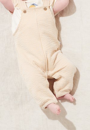 Säugling liegt und trägt beige-weiße gestreifte Latzhosen mit Knöpfen und ein weißes Shirt mit einer kleinen orangenen Grafik.