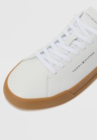 Zapatilla de cuero blanca con suela de goma, puntera redonda, cordones blancos planos y bordado de marca en el lateral. Acabado suave y acentos texturizados.