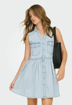 Donna che indossa un vestito di denim chiaro senza maniche con bottoni sul davanti e tasche sul petto, che tiene una borsa nera a tracolla sul braccio destro.