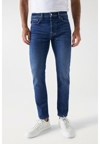 Siniset farkut slim fit -mallilla, joissa on viisi taskua, vyölenkit ja hienovarainen kuluminen. Yhdistetty valkoisiin tennareihin.