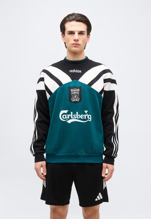 Νεαρός άντρας που φοράει ένα πετρόλ και μαύρο φούτερ ποδοσφαίρου Adidas Liverpool και μαύρο σορτς Adidas, στέκεται μπροστά σε μια απλή φόντο.