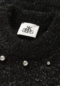 Čierny chlpatý textil s lesklými niťami, zdobený troma reflexnými perlami a bielou textíliou s čiernym logom.