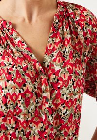 Floral-patterned top featuring vibrant red, pink, and green colors. Lightweight fabric with a v-neck and pleated details on the shoulders.