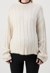 Stickad krämfärgad tröja med vertikal ribbning, hög hals och svarta kanter på ärmarna. Mjuk textur, avslappnad passform och lätt kortad design.