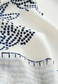 Tissu blanc avec motifs floraux et quadrillés bleus, montrant des coutures texturées et des plis doux.