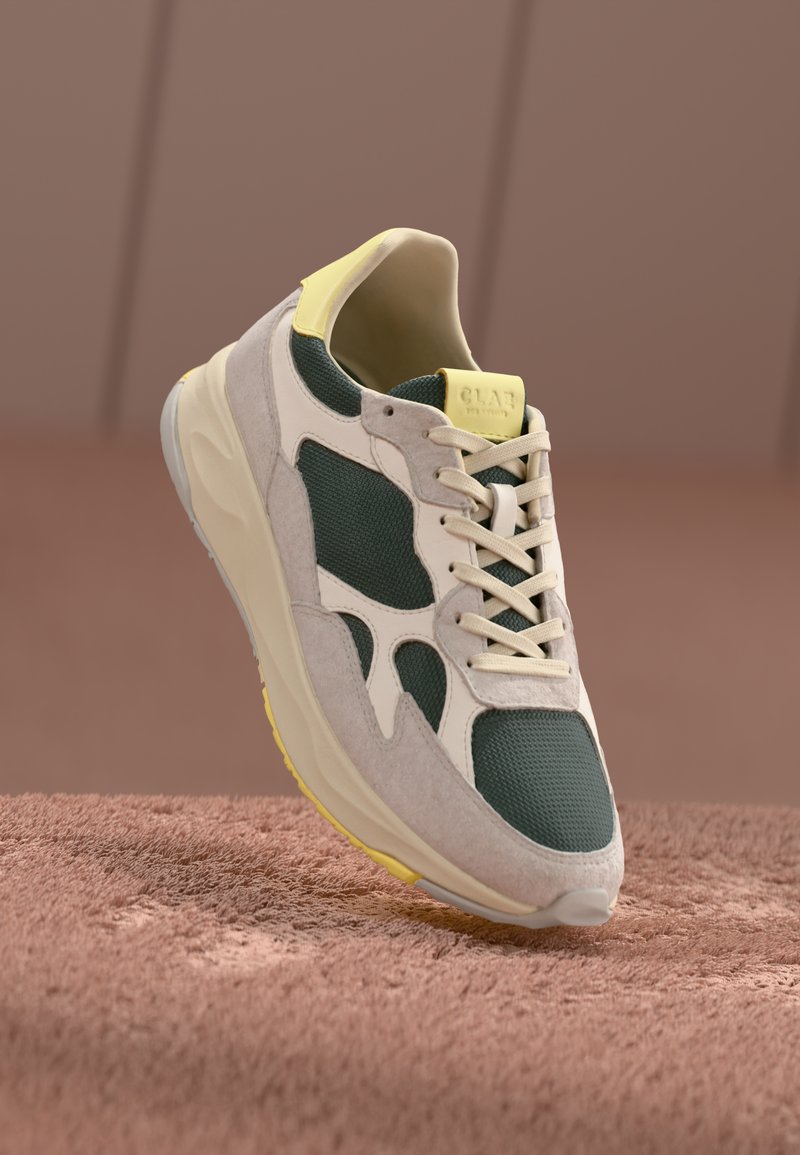 Sneaker med grønn mesh-overdel, lysegrå suede-detaljer, beige såle og gul hæltab. Har lisser og buet design for komfort.