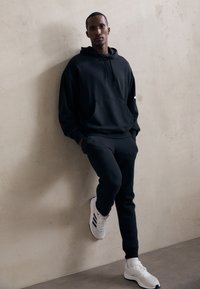 Mann lehnt an einer strukturierten Wand, trägt einen schwarzen Hoodie, schwarze Jogginghosen und weiße Sneaker mit schwarzen Streifen.