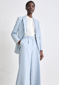 Costume sur mesure bleu clair avec un blazer à un bouton et un pantalon à jambes larges. Comprend une chemise blanche à boutons en dessous. Texture de tissu lisse.