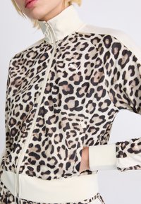 Leopard print track jacket with a high collar, cream accents, zip closure, and ribbed cuffs. Features side pockets and a soft fabric texture.
