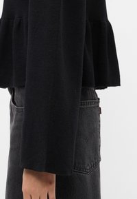 Svart strikket genser med peplumkant og lange ermer, parret med høyhansen mørk denimbukser med baklommebroderi.