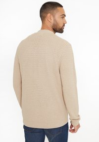 Beige gestrickter Pullover mit strukturiertem Muster, Rundhalsausschnitt und langen Ärmeln, kombiniert mit dunkelblauen Jeans.