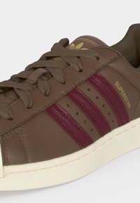 Καφέ αθλητικό παπούτσι Adidas Superstar με μαρουαν ρίγες, χρυσό λογότυπο, κορδόνια και άσπρη σόλα σε κοντινή γωνία.