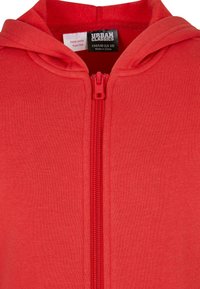 Röd zip-up hoodie i ett strukturerat tyg, med en framficka och en huva. Taggar synliga för storlek och skötselråd.