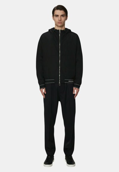 Giovane uomo in piedi, indossa una felpa nera con cerniera e sottili dettagli bianchi, pantaloni neri larghi e scarpe da ginnastica nere con suola bianca.